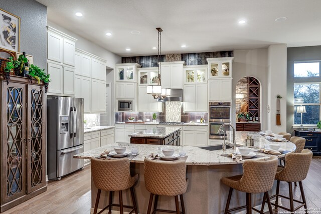 kitchen featuring light wood-type flooring, stainless steel appliances, a kitchen island with sink, light stone countertops, and wall chimney range hood