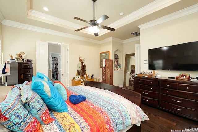 bedroom with ceiling fan, a raised ceiling, dark wood-type flooring, a walk in closet, and ornamental molding