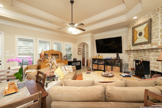 tiled living room with ceiling fan, a raised ceiling, crown molding, and a stone fireplace