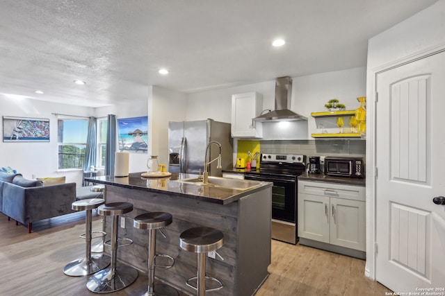 kitchen with stainless steel appliances, a kitchen breakfast bar, wall chimney range hood, an island with sink, and light hardwood / wood-style floors