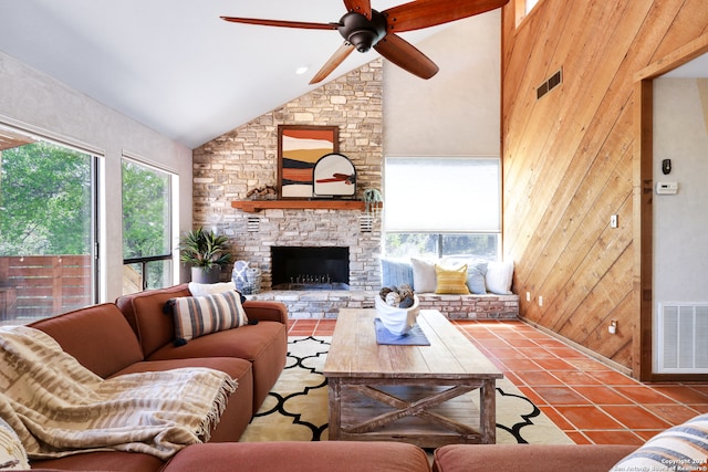 tiled living room with a stone fireplace, ceiling fan, wooden walls, and high vaulted ceiling