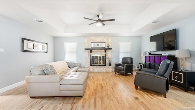 living room featuring light wood-type flooring, a raised ceiling, a stone fireplace, and ceiling fan