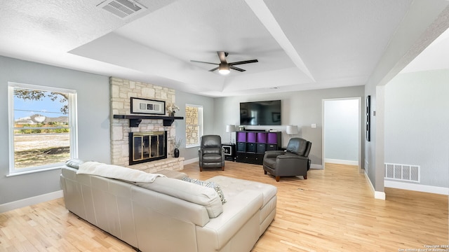 living room with a raised ceiling, a stone fireplace, a textured ceiling, ceiling fan, and light hardwood / wood-style flooring