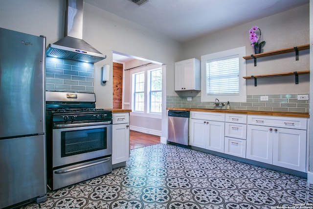 kitchen featuring wooden counters, stainless steel appliances, white cabinets, backsplash, and wall chimney exhaust hood