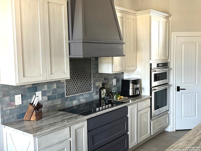 kitchen featuring light stone counters, premium range hood, black electric stovetop, stainless steel double oven, and decorative backsplash