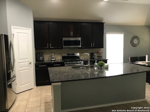 kitchen featuring a center island with sink, light tile patterned floors, appliances with stainless steel finishes, dark stone counters, and sink