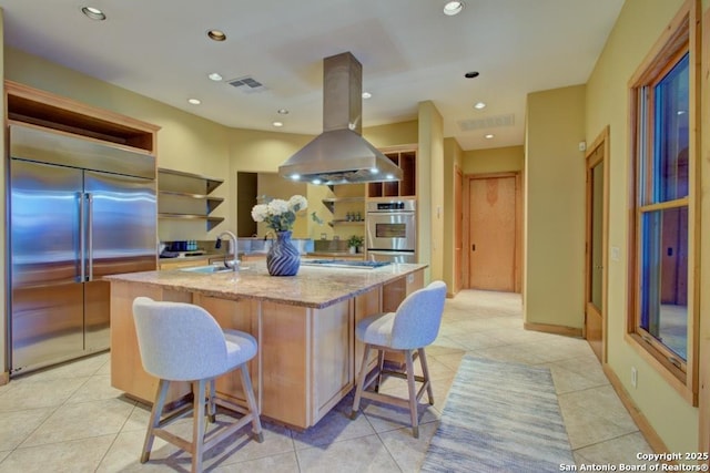 kitchen featuring appliances with stainless steel finishes, sink, island range hood, light stone countertops, and a kitchen island with sink