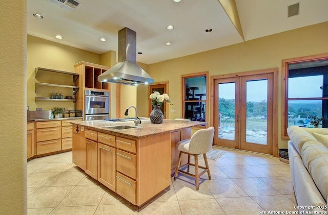 kitchen with a center island with sink, double oven, sink, island range hood, and light brown cabinetry