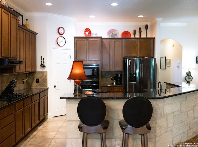 kitchen featuring dark stone counters, black appliances, light tile patterned floors, backsplash, and ornamental molding