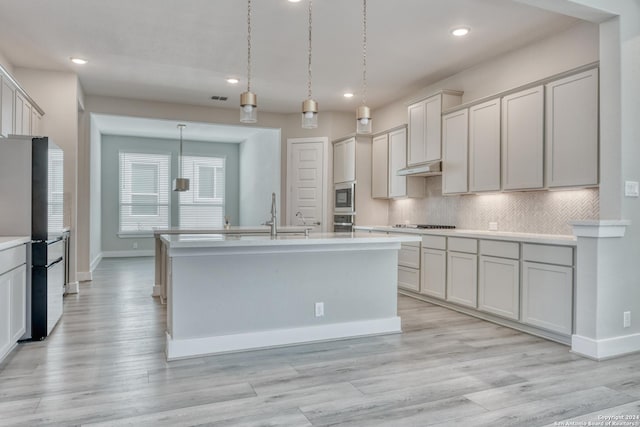 kitchen featuring light wood-style flooring, light countertops, appliances with stainless steel finishes, backsplash, and a center island with sink