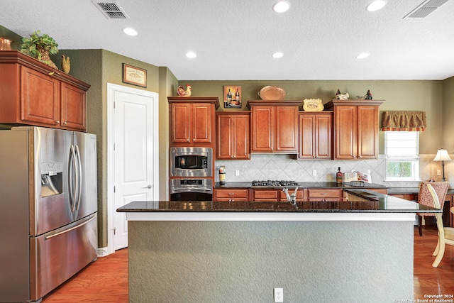 kitchen featuring appliances with stainless steel finishes, light hardwood / wood-style floors, tasteful backsplash, a kitchen island, and dark stone countertops