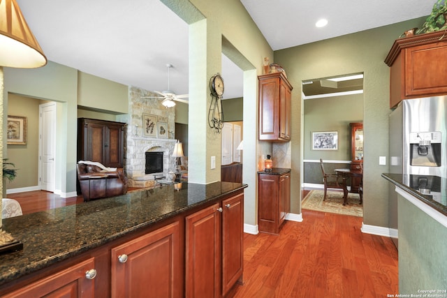 kitchen featuring dark stone countertops, stainless steel refrigerator with ice dispenser, wood-type flooring, ceiling fan, and a stone fireplace