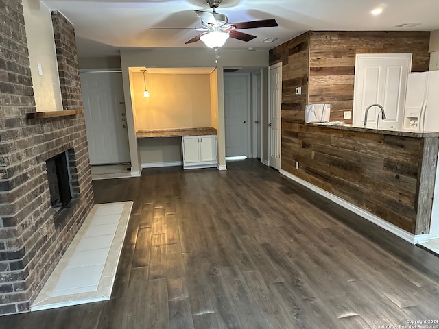 unfurnished living room with sink, wooden walls, dark wood-type flooring, ceiling fan, and a brick fireplace