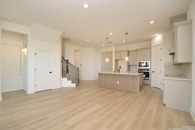 kitchen with a center island with sink, built in microwave, hanging light fixtures, stainless steel oven, and light wood-type flooring