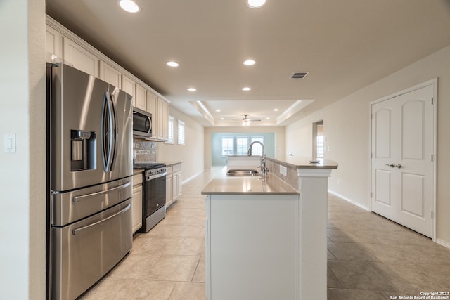 kitchen with light tile patterned flooring, sink, a kitchen island with sink, a tray ceiling, and stainless steel appliances