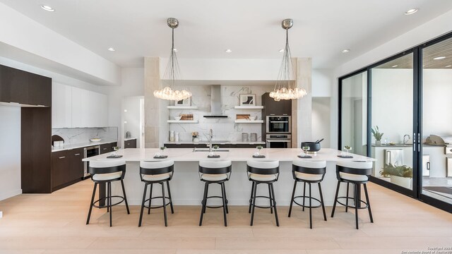 kitchen featuring dark brown cabinets, tasteful backsplash, white cabinetry, a center island with sink, and wall chimney range hood