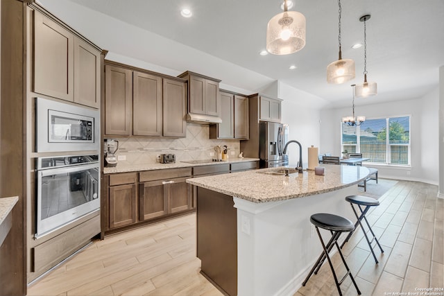 kitchen with backsplash, decorative light fixtures, appliances with stainless steel finishes, light stone countertops, and an island with sink