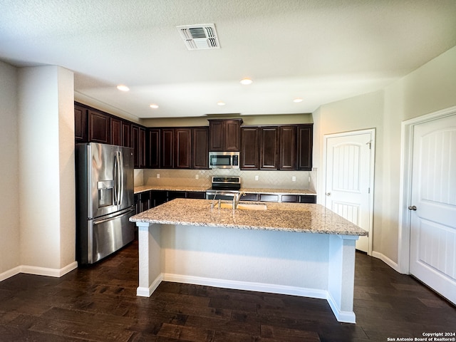 kitchen featuring dark brown cabinetry, dark wood-type flooring, an island with sink, appliances with stainless steel finishes, and light stone countertops