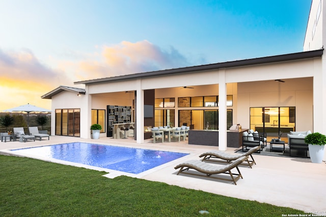 pool at dusk with ceiling fan, an outdoor hangout area, a patio area, and a yard