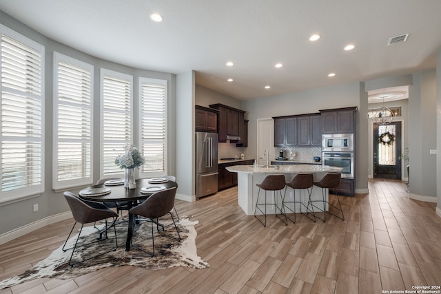 kitchen featuring appliances with stainless steel finishes, backsplash, dark brown cabinets, a center island with sink, and light wood-type flooring