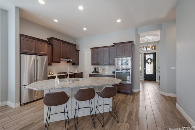 kitchen with backsplash, dark brown cabinetry, light stone countertops, a center island with sink, and stainless steel appliances