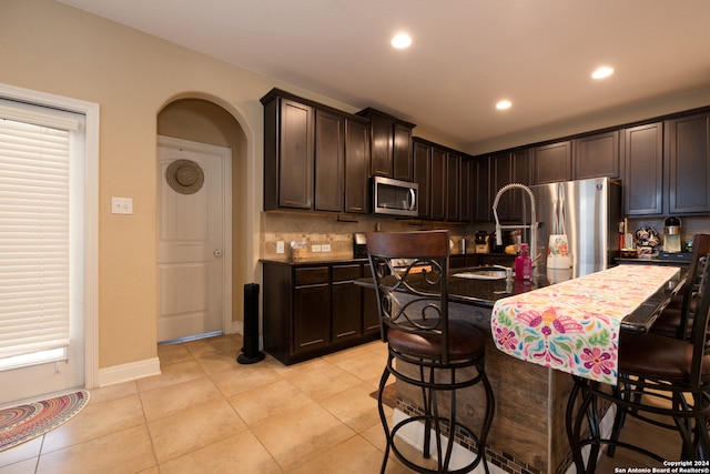 kitchen with light tile patterned flooring, an island with sink, a breakfast bar, stainless steel appliances, and dark brown cabinets