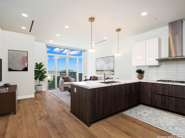 kitchen featuring hanging light fixtures, light hardwood / wood-style floors, white cabinetry, sink, and wall chimney range hood