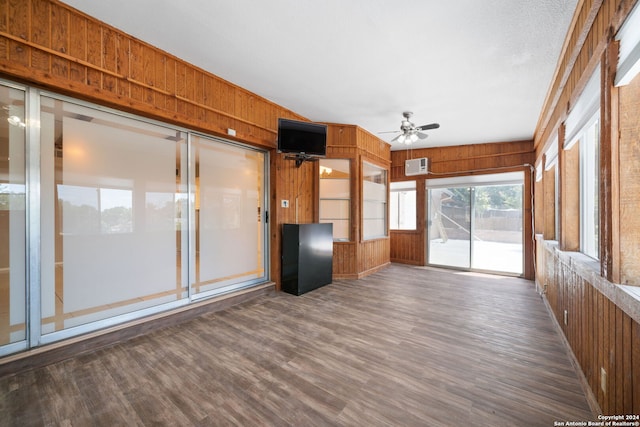 unfurnished living room featuring dark wood-style floors, ceiling fan, wooden walls, and a sunroom