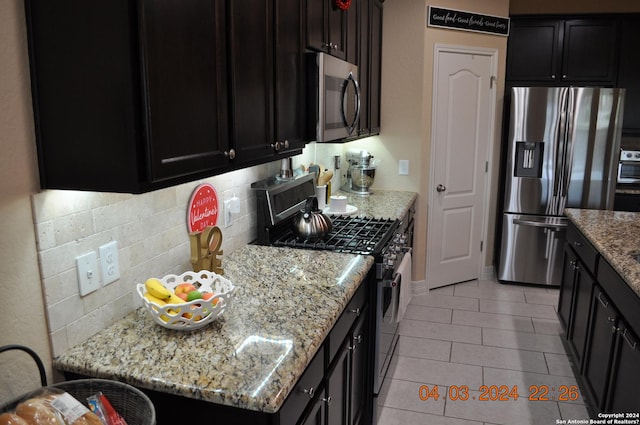 kitchen with light tile patterned floors, dark cabinets, appliances with stainless steel finishes, light stone countertops, and tasteful backsplash