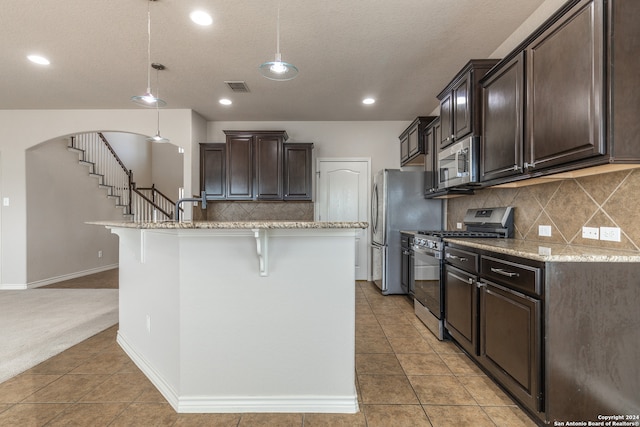 kitchen featuring an island with sink, stainless steel appliances, dark brown cabinets, a breakfast bar, and decorative light fixtures