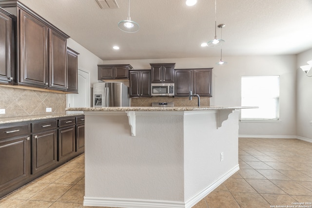 kitchen with decorative light fixtures, stainless steel appliances, dark brown cabinets, and an island with sink