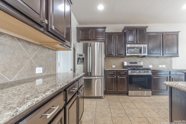 kitchen featuring dark brown cabinets, stainless steel appliances, light stone countertops, light tile patterned floors, and tasteful backsplash