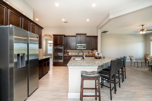 kitchen with backsplash, stainless steel appliances, light hardwood / wood-style flooring, light stone counters, and a kitchen island with sink