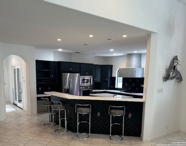 kitchen featuring backsplash, stainless steel appliances, wall chimney exhaust hood, kitchen peninsula, and a breakfast bar area