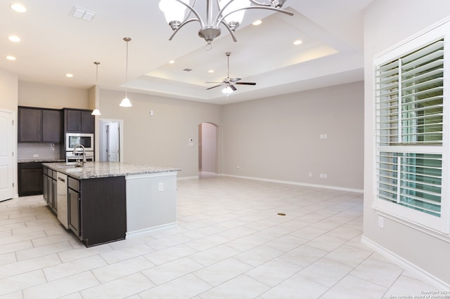 kitchen featuring ceiling fan with notable chandelier, a center island with sink, a raised ceiling, decorative light fixtures, and light stone countertops