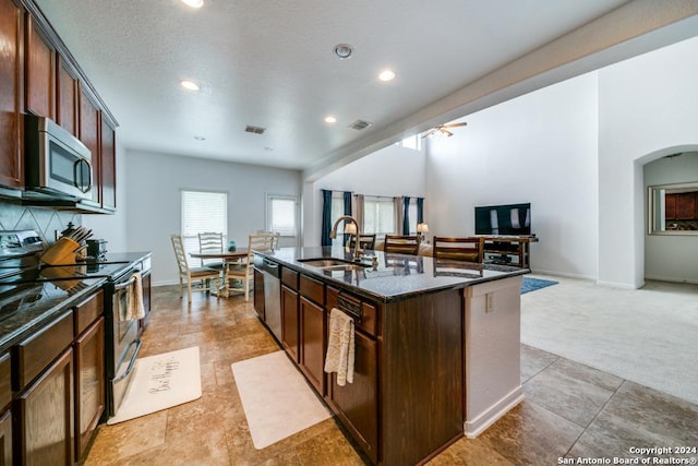 kitchen featuring arched walkways, appliances with stainless steel finishes, a sink, an island with sink, and dark stone counters