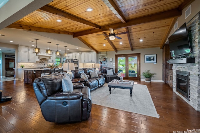 living room with beamed ceiling, a stone fireplace, dark wood-type flooring, and ceiling fan