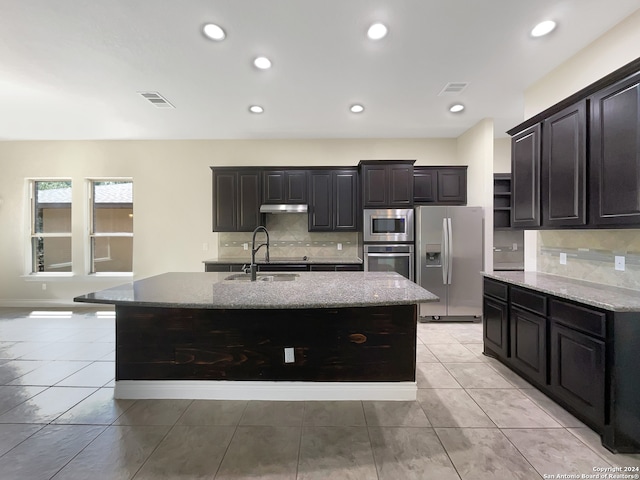 kitchen featuring appliances with stainless steel finishes, backsplash, an island with sink, and light stone countertops