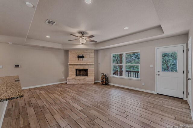 unfurnished living room featuring a tray ceiling, a stone fireplace, ceiling fan, and light hardwood / wood-style floors