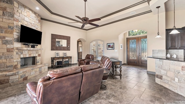 living room featuring dark tile patterned floors, crown molding, a raised ceiling, a stone fireplace, and ceiling fan