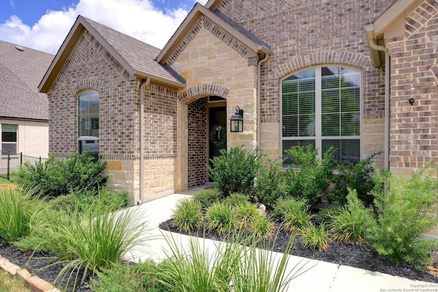 view of exterior entry featuring stone siding, brick siding, a shingled roof, and fence