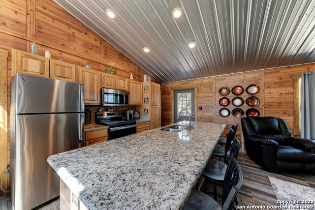 kitchen featuring wood walls, stainless steel appliances, a center island with sink, and vaulted ceiling