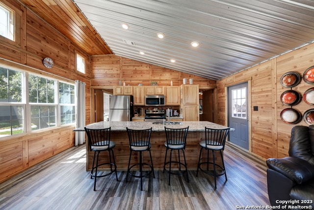 kitchen with wooden walls, stainless steel appliances, lofted ceiling, wood-type flooring, and an island with sink