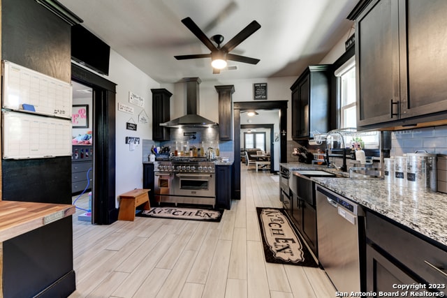 kitchen with stainless steel appliances, light stone counters, ceiling fan, wall chimney exhaust hood, and light hardwood / wood-style floors