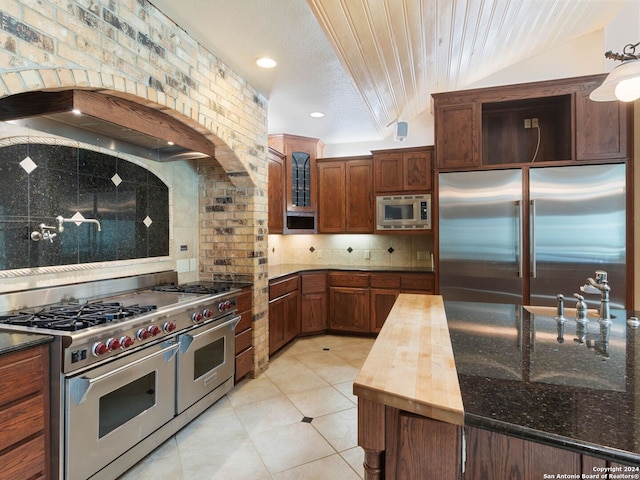 kitchen with custom exhaust hood, built in appliances, light tile patterned floors, glass insert cabinets, and backsplash