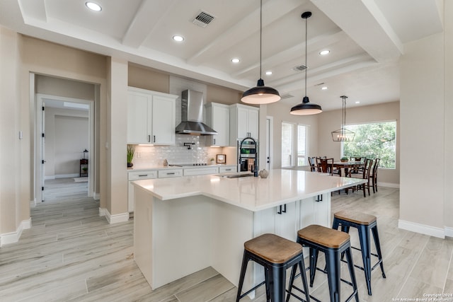 kitchen featuring wall chimney exhaust hood, light hardwood / wood-style flooring, pendant lighting, a kitchen island with sink, and white cabinetry