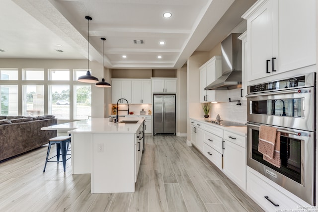 kitchen featuring wall chimney exhaust hood, a center island with sink, light hardwood / wood-style flooring, appliances with stainless steel finishes, and white cabinets