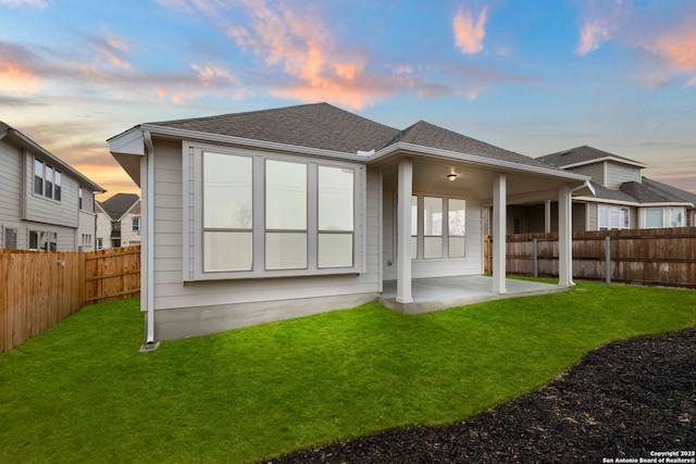 back of house with a fenced backyard, a shingled roof, a lawn, and a patio