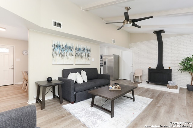 living room featuring light wood-type flooring, a wood stove, ceiling fan, lofted ceiling with beams, and brick wall