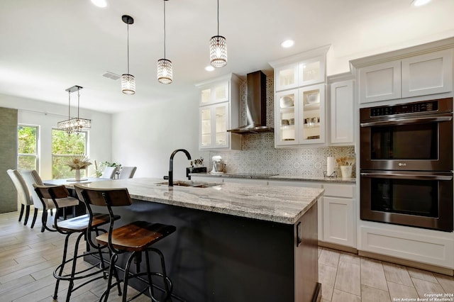 kitchen with wall chimney exhaust hood, an island with sink, hanging light fixtures, sink, and double oven